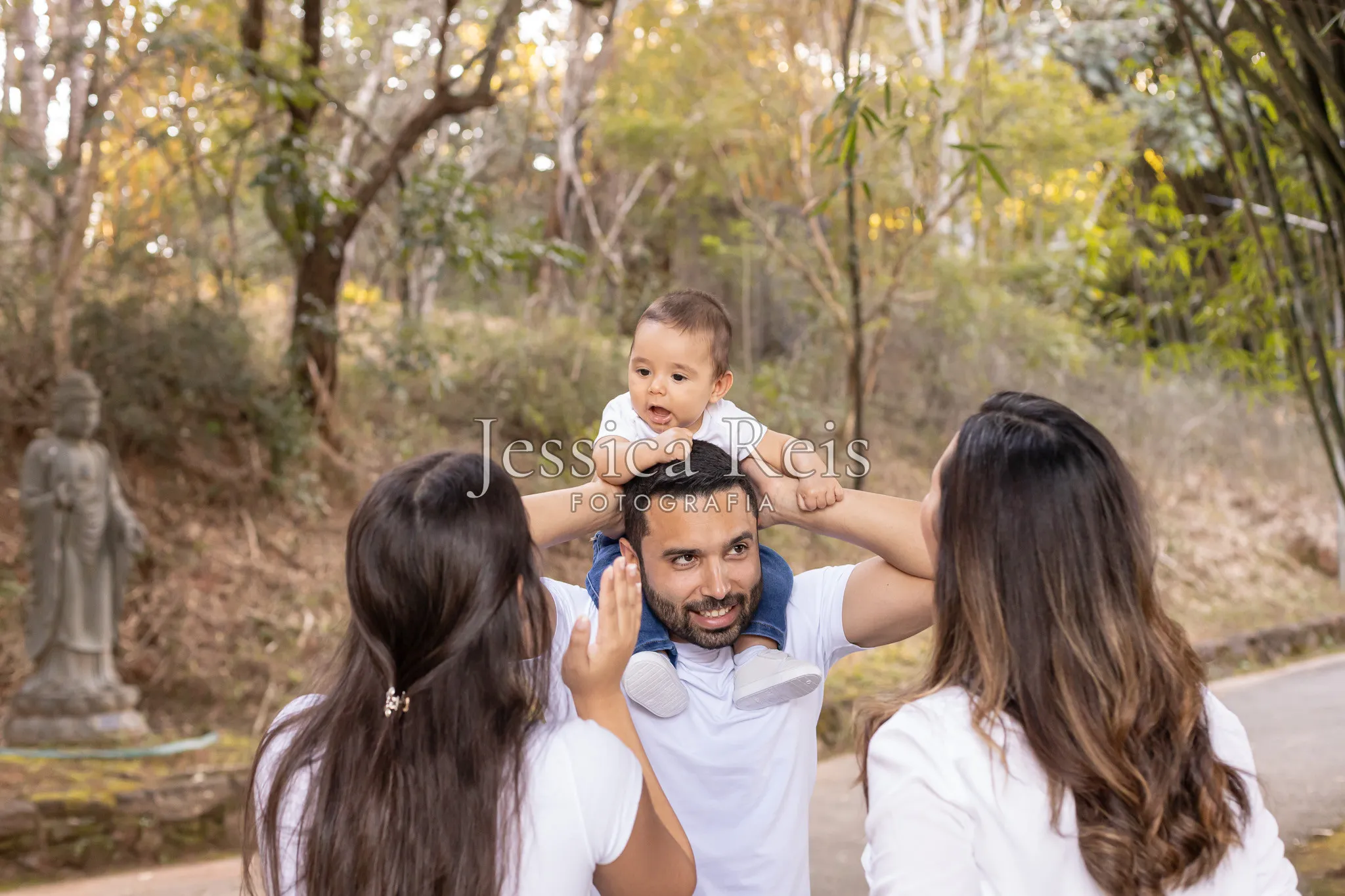 Bebê no colo do pai durante passeio em família no parque equanto a mãe e irmã mais velha brincam com o bebê